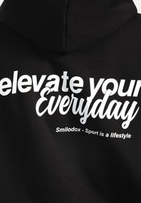 Hoodie nera con una texture morbida, con testo bianco stampato: "eleva il tuo quotidiano" e "Smilodox - Lo sport è uno stile di vita" sulla schiena.