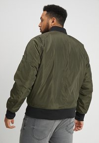 Giubbotto bomber verde oliva con una texture liscia, polsini e orlo a coste, e tasche laterali. Presenta un colletto nero e dettagli di cucitura sottili.