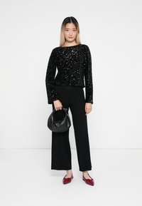 Top lung cu mânecă lungă, decorat cu paiete negre, asortat cu pantaloni negri cu croială largă, balerini roșii cu vârf ascuțit și o geantă rotundă neagră.