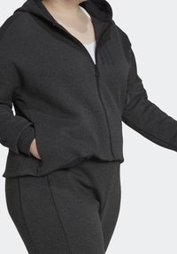 Personne portant un sweat à capuche gris foncé zippé et un pantalon assorti, une main dans la poche, devant un fond uni.