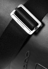 Detail einer schwarzen Lederhandtasche mit einem strukturierten schwarzen Riemen und einer silbernen Metallschnalle. Die Schnalle hat eine eingravierte Markierung mit dem Schriftzug "Piquadro."