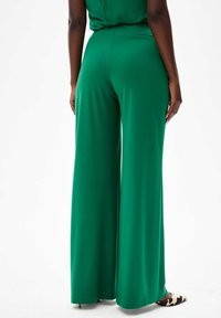 Groene wijde broek van soepel materiaal, met een hoge taille en een uitlopende silhouet. De zoom is schoon en schoenen met een gemustreerd accent zijn zichtbaar.