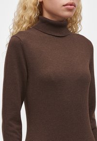 Femme aux cheveux blonds bouclés portant un pull à col roulé marron ajusté, montrée des épaules aux lèvres sur un fond uni.