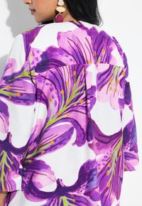 Løse bluser med store lilla blomster mønstre på hvid baggrund, med grønne detaljer og en rygflæse.