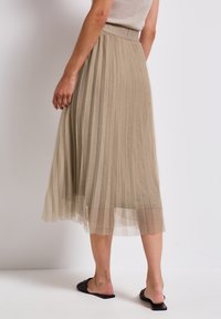 Keskikokoisen vaaleanbeige tüllihameen liehukerroksilla, jossa on korkea vyötärö ja virtaava siluetti. Yhdistetty mustiin liukusandaaleihin.