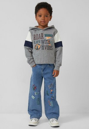 Sweat-shirt à capuche gris avec des textes graphiques et des designs, associé à un jean bleu avec des patchs colorés. Des baskets blanches complètent la tenue.