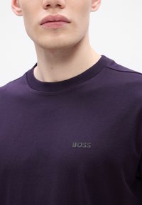 Κοντινό πλάνο ενός ατόμου που φοράει ένα σκούρο μωβ μπλουζάκι με λαιμόκοψη, με ένα μικρό ασημένιο λογότυπο "BOSS" στο αριστερό στήθος.