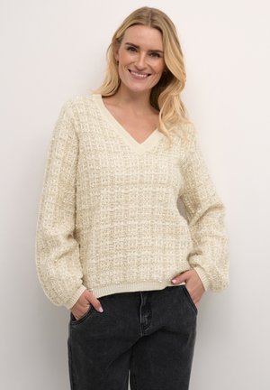 Beige pletený svetr s výstřihem do V, strukturovaným vzorem a dlouhými rukávy. Spárován s tmavě šedými kalhotami, ruce zasunuté v kapsách.