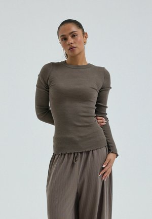 Kvinde med trukket hår tilbage iført en tætsiddende langærmet ribbet taupe-top og løstsiddende matchende teksturerede bukser, poserer mod en ensfarvet grå baggrund.