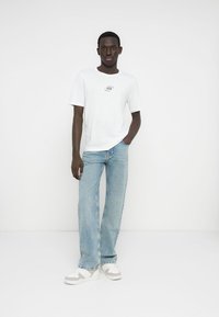 Biała bawełniana koszulka z niewielkim nadrukiem na piersi, w połączeniu z luźnymi jasnoniebieskimi jeansami i białymi trampkami z jasnymi akcentami.