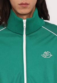 Grøn zip-up jakke med høj krave, hvid lynlås og lille broderet logo på venstre bryst. Glat stof med ribbet krave.