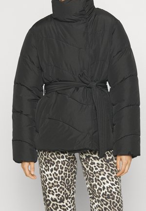 Persoon draagt een zwart gewatteerd jasje met een gestrikte riem in de taille en een broek met luipaardprint tegen een effen achtergrond.
