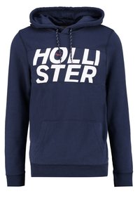 Sudadera azul marino de tejido suave, con un gran logo blanco "HOLLISTER" en el pecho, cordones y un bolsillo delantero.