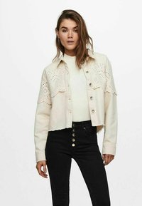 Veste en denim beige coupée avec un motif floral découpé au laser et des boutons argentés, portée sur un haut blanc, associée à un pantalon noir taille haute.