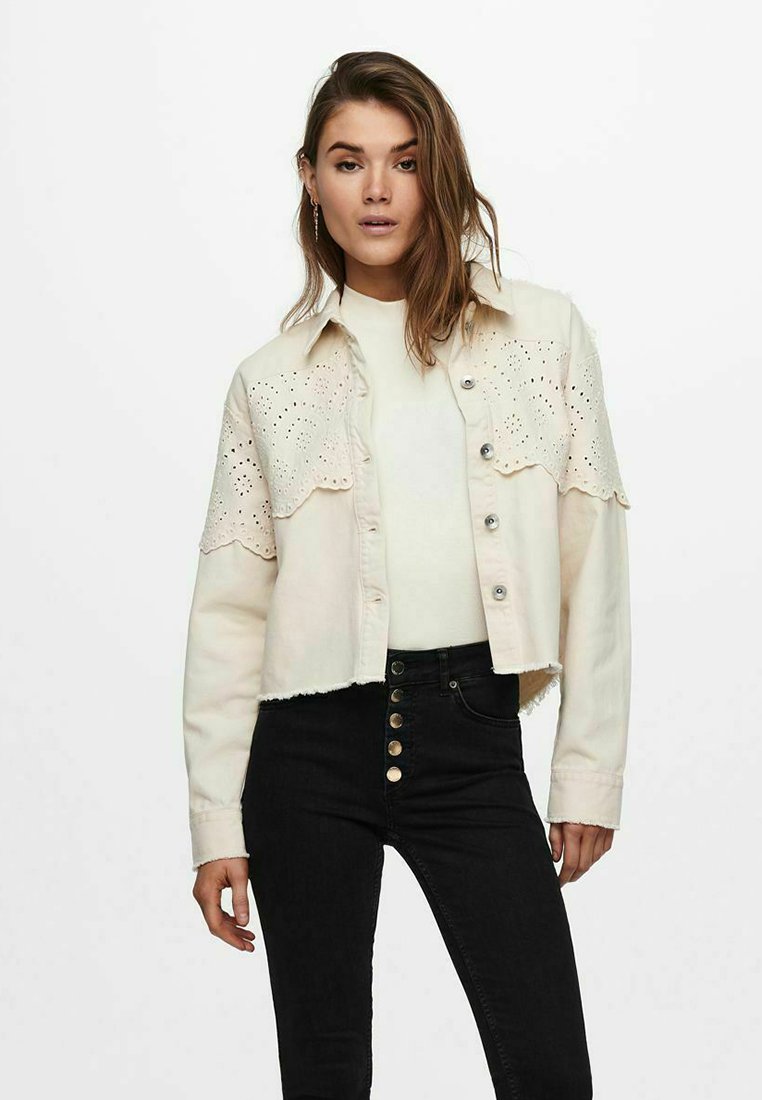 Veste en denim beige coupée avec un motif floral découpé au laser et des boutons argentés, portée sur un haut blanc, associée à un pantalon noir taille haute.
