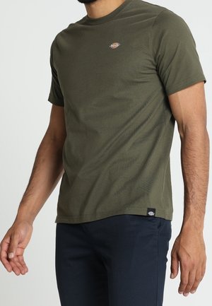 Homme portant un t-shirt à manches courtes vert olive avec un petit logo sur la poitrine et un pantalon noir, debout devant un fond clair uni.