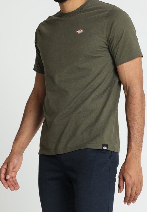 Homme portant un t-shirt à manches courtes vert olive avec un petit logo sur la poitrine et un pantalon noir, debout devant un fond clair uni.