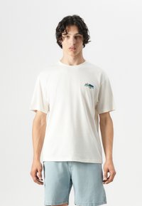 Cream-colored cotton t-shirt with short sleeves featuring a small colorful printed logo on the left chest area. Paired with light blue shorts.