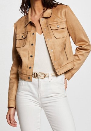 Vrouw draagt een tan suède cropped jack over een witte top, gecombineerd met witte hoge taillebroeken en een witte riem met gouden gesp.