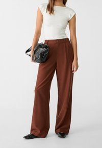 Wijde broek in bruine stof, met een hoge taille en een knoopsluiting, gecombineerd met een aansluitende witte top en een zwarte handtas.
