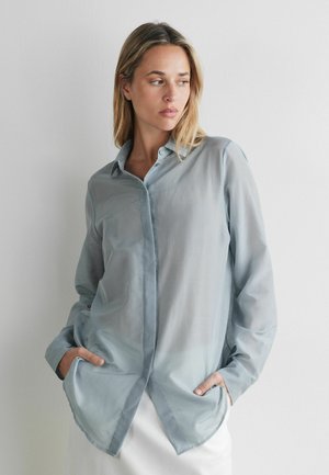 Frau mit blondem Haar, die ein hellblaues, langärmliges Hemd mit Knöpfen trägt, steht vor einer weißen Wand mit den Händen in den Taschen.