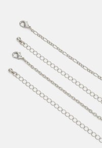 Srebrne metalowe łańcuchy o różnych kształtach ogniw, w tym owalnych i okrągłych. Zawierają zapięcie typu homar i gładki kulisty koniec. Minimalistyczny design.