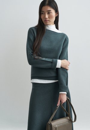 Suéter de punto en color azul verdoso con un acabado texturizado, superpuesto a un cuello alto blanco, combinado con una falda a juego y llevando un bolso marrón.