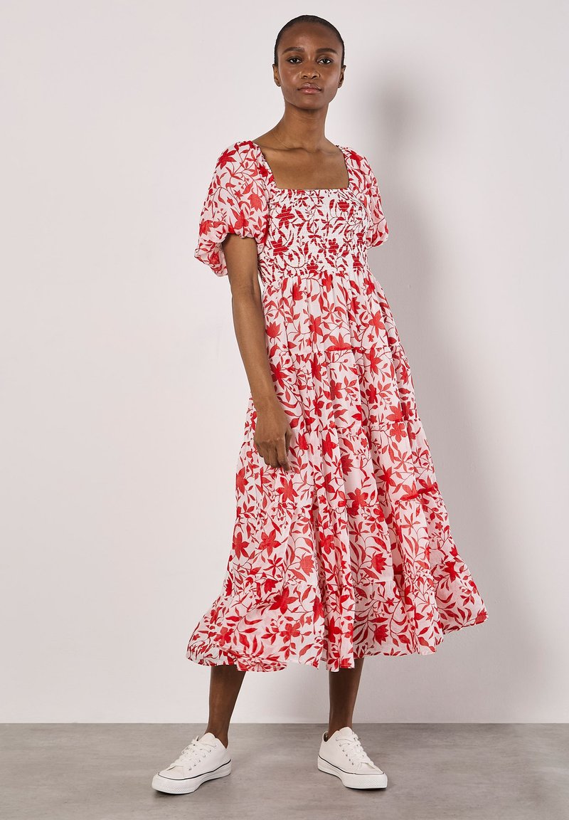 Rotes Blumen-Midi-Kleid mit Puffärmeln, quadratischem Ausschnitt und gerafftem Oberteil. Hergestellt aus leichtem Stoff, mit einem volantranden Saum. Kombiniert mit weißen Sneakern.