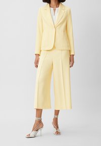 Blazer ajusté jaune et pantalon évasé, fabriqués en tissu lisse. Les chaussures sont des escarpins à brides chevilles blancs. Design minimaliste, sans motifs.