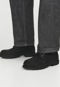 Botas de tobillo de ante negro con suela de goma, con ojetes de metal y cordones, combinadas con pantalones de mezclilla oscuros con costuras en contraste.