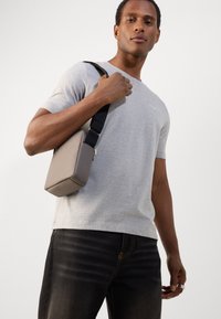 Tricou gri din bumbac, purtat cu blugi închiși la culoare. Ține în mână o geantă mică, rectangulară, bej, cu o curea neagră. Textură netedă, design minimalist.
