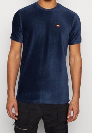Mand iført en marineblå kortærmet fløjls-T-shirt med et lille orange og rødt logo på brystet og sorte cargobukser.