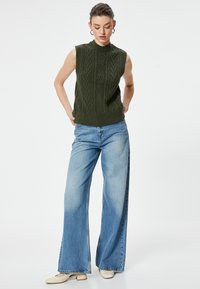 Suéter sin mangas color caqui de punto con patrón de cable texturizado, combinado con jeans de pierna ancha en azul claro y zapatos de tacón bloc en beige.