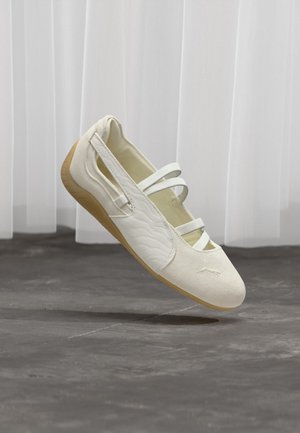 Elfenbeinfarbener Mary-Jane-Schuh mit drei Riemen, strukturierten Seiten und hellbrauter flexibler Sohle, schwebend über grauem Boden.