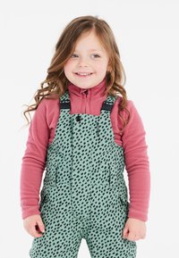 Salopette verde a fantasia con forme nere, spalline regolabili e tasche frontali, indossata sopra un top in pile rosa con zip.