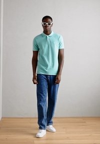 Polo vert menthe, jean bleu et baskets blanches. Modèle portant des lunettes de soleil blanches, se tenant sur un sol en bois contre un mur neutre.