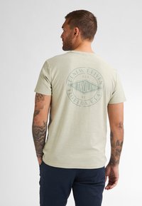 Světle zelené bavlněné tričko s kruhovou grafikou "Petrolind" na zadní straně, obsahující text a vintage design. Krátké rukávy a volný střih.