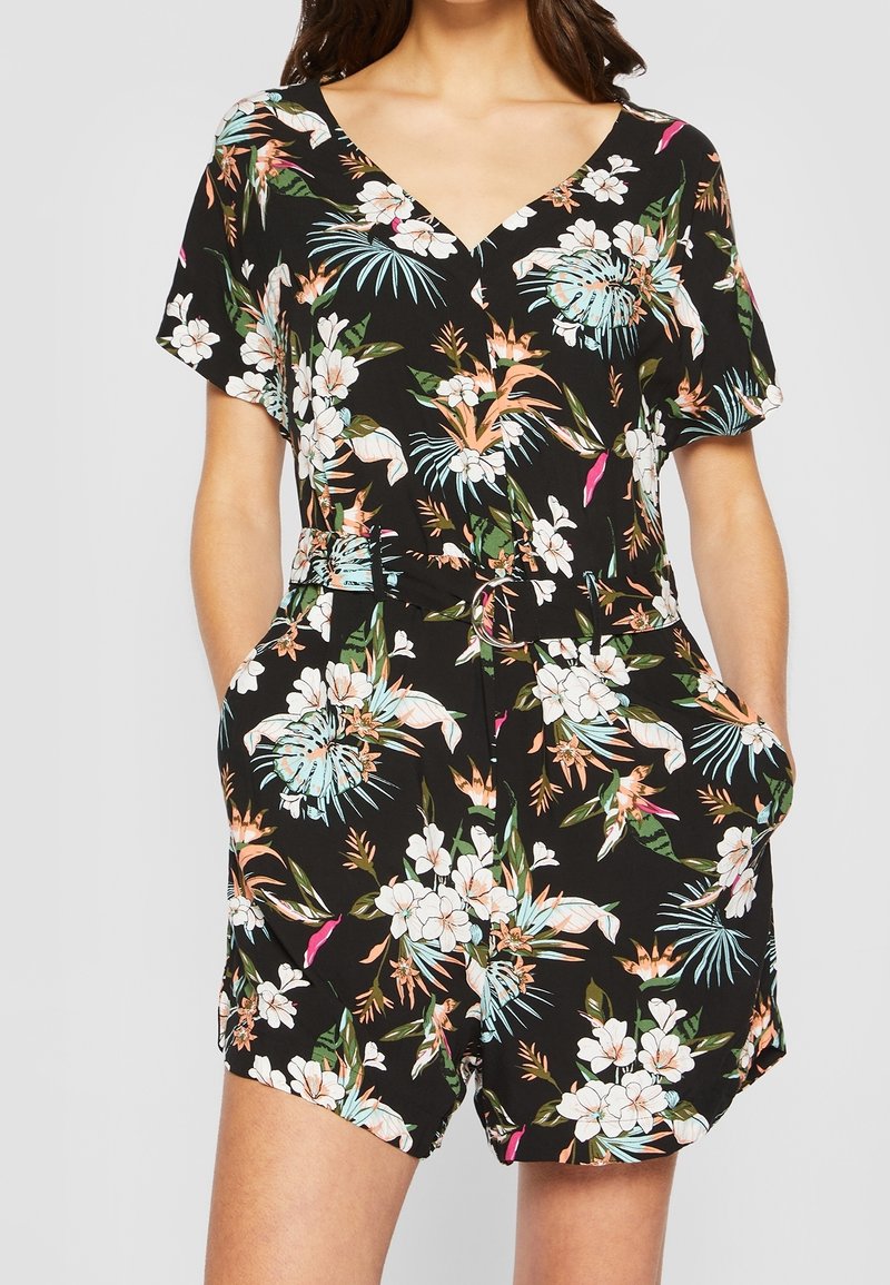 Femme portant une combinaison courte noire à manches courtes avec un motif floral tropical coloré et des poches, nouée à la taille.