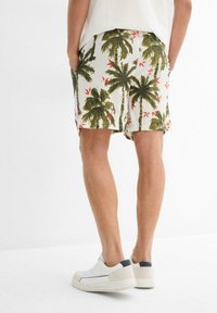 Shorts pour hommes avec une base blanche ornée d'un imprimé de palmiers verts et de fleurs rouges, avec des poches latérales et une coupe décontractée. Associés à des baskets blanches.