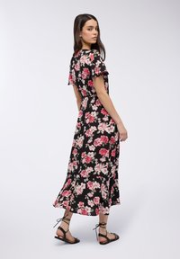 Donna in un vestito midi floreale nero con maniche corte, che guarda oltre la sua spalla, indossando sandali neri allacciati su uno sfondo neutro.