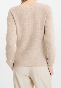 Femme portant un pull beige en maille et un pantalon couleur crème, vue de dos sur un fond blanc.