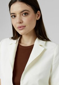 Blazer blanc avec un col cranté, porté sur un haut côtelé marron. Texture lisse, coupe ajustée et coutures subtiles accentuent le design.