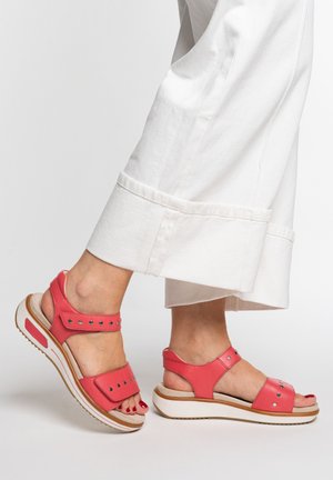 Feet wearing red open-toe platform sandals with ankle straps paired with white wide-leg cropped pants against a plain background.