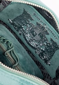 L'interno della borsa in velluto turchese presenta un logo impresso e un design intricato. La cerniera con catena decorativa e il rivestimento a fantasia scura contrastano con la morbidezza del tessuto.