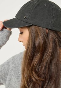 Mörkgrå bomull baseballkeps med en böjd skärm, prydd med en liten metallaccent, bärs lätt lutad, mot en grå tröjbakgrund.