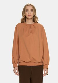 Przesadnie duża, rdzawa bluza w miękkim materiale z zebranym dekoltem i elastycznymi mankietami, w połączeniu z brązowymi spodniami.
