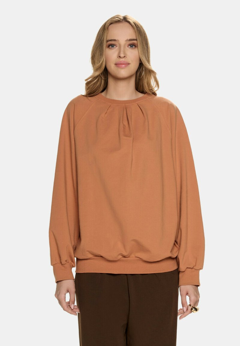 Przesadnie duża, rdzawa bluza w miękkim materiale z zebranym dekoltem i elastycznymi mankietami, w połączeniu z brązowymi spodniami.