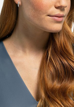 Nahaufnahme vom Hals und unteren Gesicht einer Frau mit langen roten Haaren, die silberne Creolen und ein blaues V-Ausschnitt-Oberteil trägt.