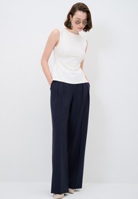 Weißes ärmelloses Top mit einem Peplum-Taille und navyblauen weiten Hosen. Das Modell trägt Sonnenbrillen und beige High Heels. Einfaches Design, glatte Textur.