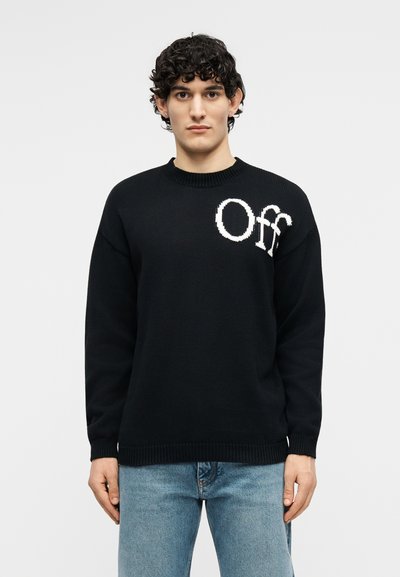 Νεαρός άνδρας με σγουρά μαλλιά που φοράει μαύρο πουλόβερ με τη λέξη «Off» σε λευκό στην αριστερή πλευρά, συνδυασμένο με ανοιχτόχρωμα μπλε τζιν, στέκεται με το πρόσωπο προς τα εμπρός.